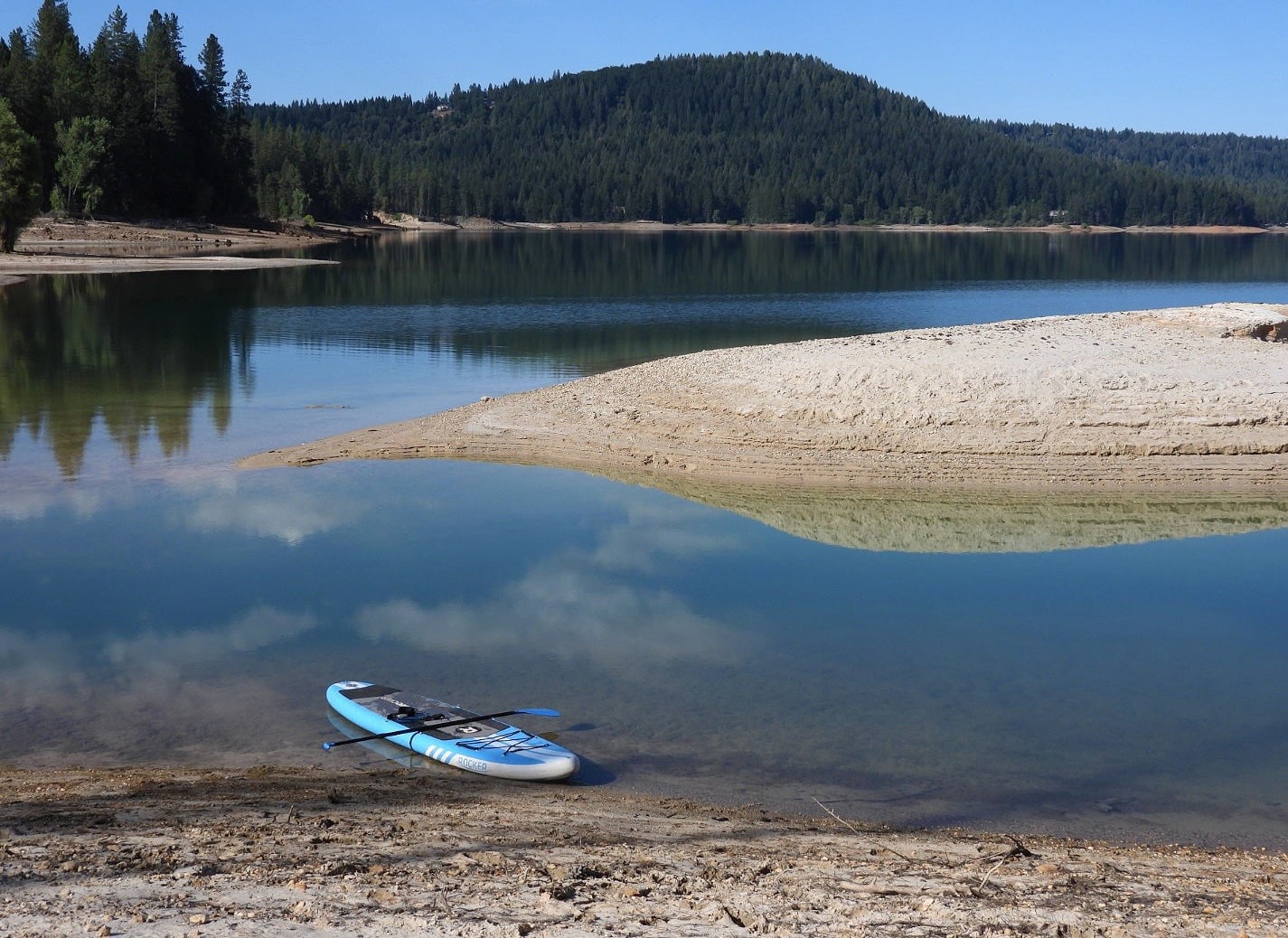 A stand-up paddleboard rests on a Sierra Nevada lake shore, pine-covered hills reflected in calm water.