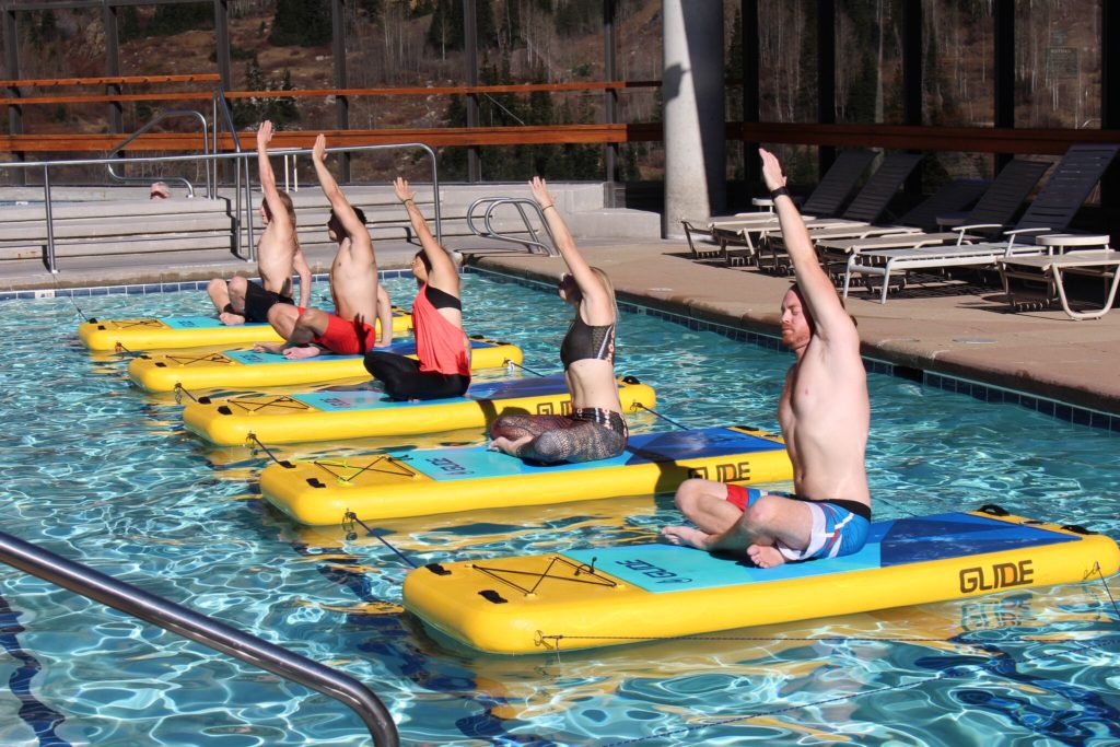 A row of students kneels on yellow Glide pool-fit boards with one arm raised, sunlit mountain backdrop visible through the open pool deck.